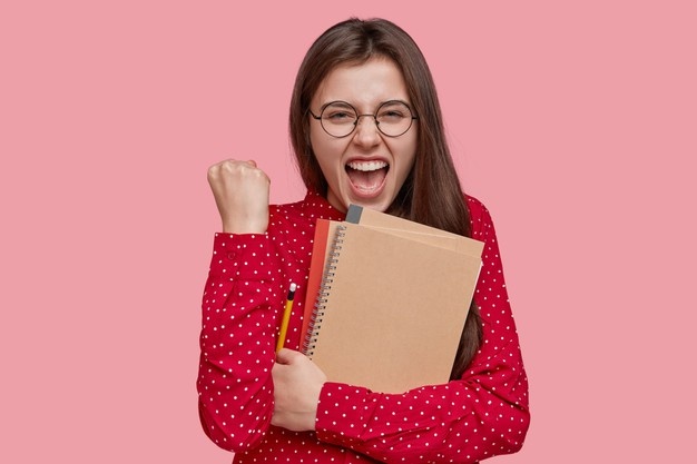 positive-happy-teacher-clenches-fist-with-joyful-expression-holds-spiral-notepad-writes-notes-notepad-dressed-red-shirt_273609-25182